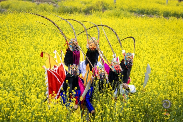 $vo['fname']|default='春日花田，地戏表演者穿行其间。镜头捕捉彩衣与花海交映，脸谱在光影中忽明忽暗。定格转身时衣袂拂过花枝的瞬间，让六百年戏韵与自然生机，在快门声里共舞。